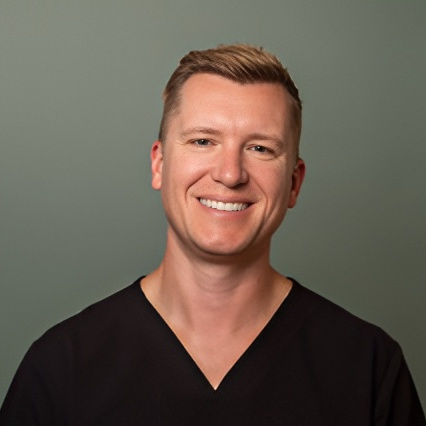 Professional headshot of a smiling male dental team member in black scrubs against a neutral background.