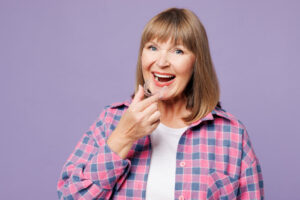 Elderly happy blonde woman 50s years old wear violet shirt casual clothes hold in hand invisible transparent aligners, invisalign dental braces isolated on plain purple background. Lifestyle concept